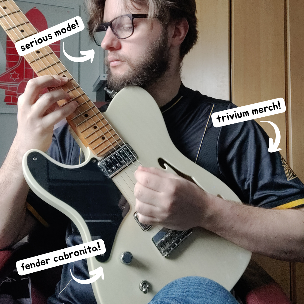 Luke sat at a desk playing a Fender Cabronita guitar. There are 3 popouts. One points to Luke's face and says 'serious mode!'. Another points to Luke's t shirt and says 'trivium merch!'. The last one points to Luke's guitar and says 'Fender Cabronita!'